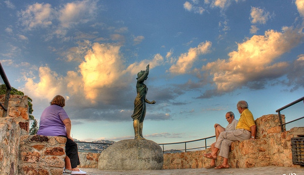 Un grupo de visitante descansa relajado junto al mar frente a la estatua de la Mujer Marinera (Dona Marinera) de Lloret de Mar, Costa Brava